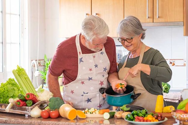 Smiling couple of seniors in chefs aprons prepare vegetables for a soup together in the home kitchen.