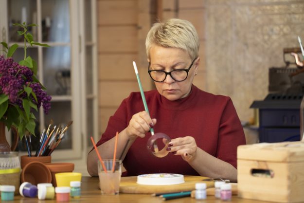 keen middle-aged woman paints wooden bracelet in country house. hobby of drawing. The Reasons Why You Need Creative Spaces As You Age In Retirement Homes In Vinton, VA