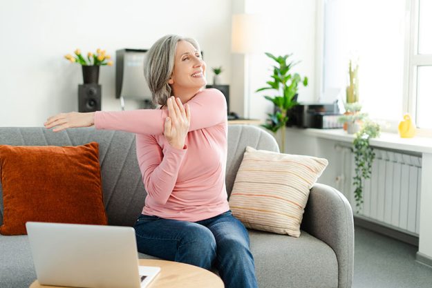Smiling active senior woman stretching arms, doing exercises sitting on comfortable sofa relaxing at home. Happy female, freelancer taking a break, resting. Natural beauty, healthy lifestyle concept