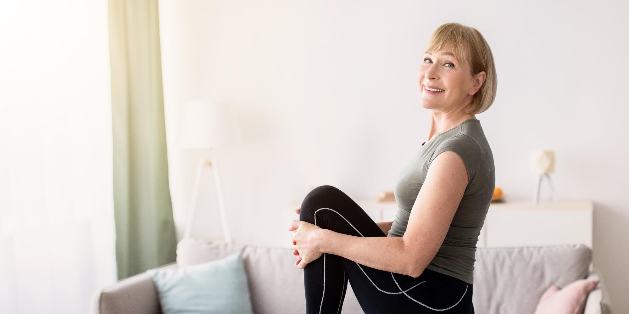 Sporty senior woman stretching her leg and smiling at camera indoors, free space. Domestic trainings during coronavirus